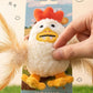 A hand holds a plush chicken-shaped pet toy attached to a printed backing card. The toy has a white fuzzy body, orange comb, yellow beak and feet, and attached feather wings.